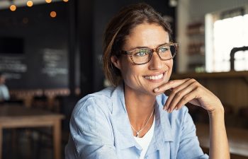 Woman with glasses smiling indoors, seated at a table with a hand under her chin, wearing a light blue shirt. Background features blurred furniture and soft string lights.