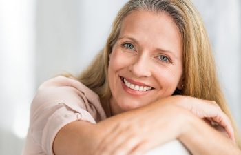 A woman with long blonde hair smiles, resting her arms on a surface, wearing a light-colored shirt.